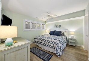 Une chambre à coucher comprenant un lit, un ventilateur de plafond, une fenêtre avec des volets, une table de chevet avec une lampe et un tapis.