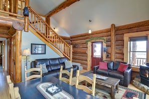 A log cabin interior with a staircase, leather sofas, wooden chairs, a dining table, and a fireplace.