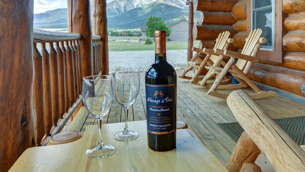 A wooden table with a bottle of wine and two glasses, set against a backdrop of mountains and a wooden cabin. - Creekside Log Cabin- conveniently located and direct creek access  (Buena Vista)