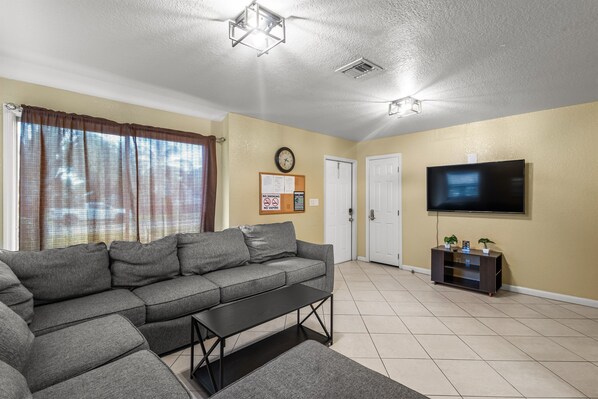 A living room with a grey sectional sofa, a small black coffee table, a flat-screen TV on a wooden stand, and a door in the background.
