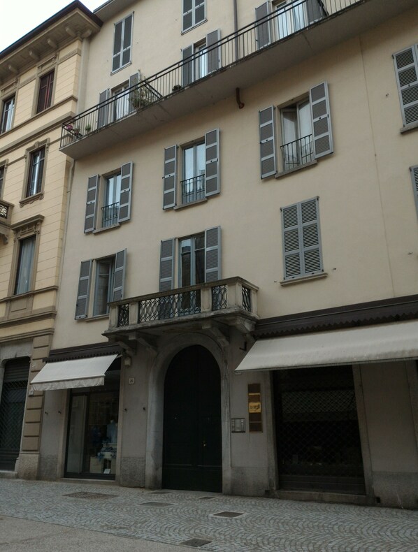 A multi-story building with a central entrance, large windows with shutters, and a balcony with metal railings.