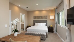 A bedroom with a bed, bedside tables, a TV, a window with curtains, a dining table with chairs, and a potted plant.