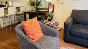 Een woonkamer met een grijze fauteuil en een blauwe bank, een eetgedeelte met een tafel en stoelen, en een keuken met houten kastjes en een fornuis.