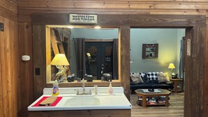Interior de una cabaña de madera con baño que incluye un lavamanos, un espejo y un cartel que dice 'SE BUSCA' para un lavavajillas.