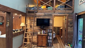 Interior rústico de una cabaña con cama en altillo, escalera de madera, televisor, estantería y un cartel que dice "BURCH CHAPEL PL".