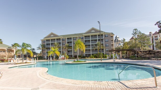 A swimming pool with a brick deck, surrounded by palm trees and a multi-story hotel building.