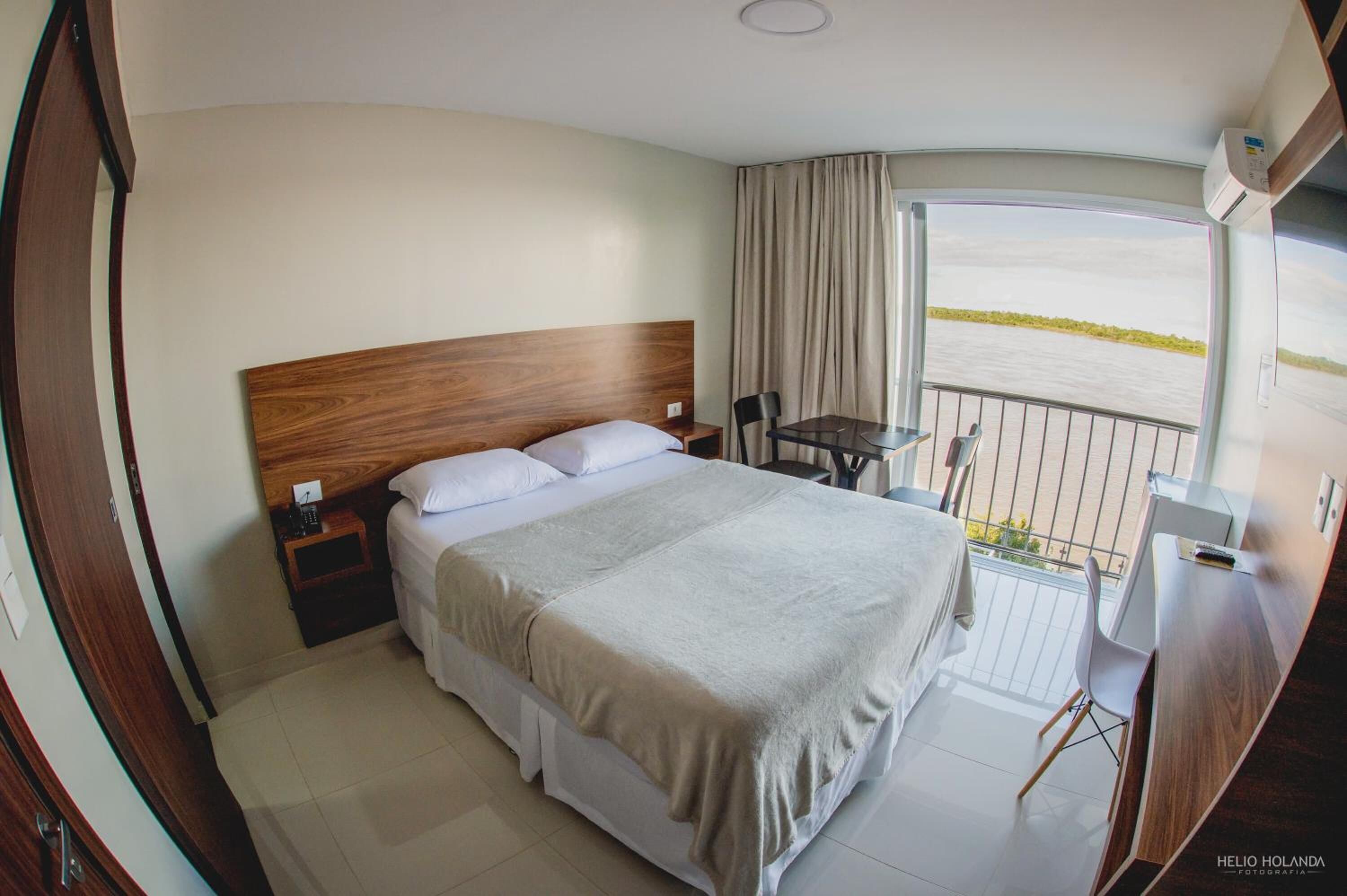 A bedroom with a large bed, a bedside table, a chair, a small table, and a balcony with a view of the water.