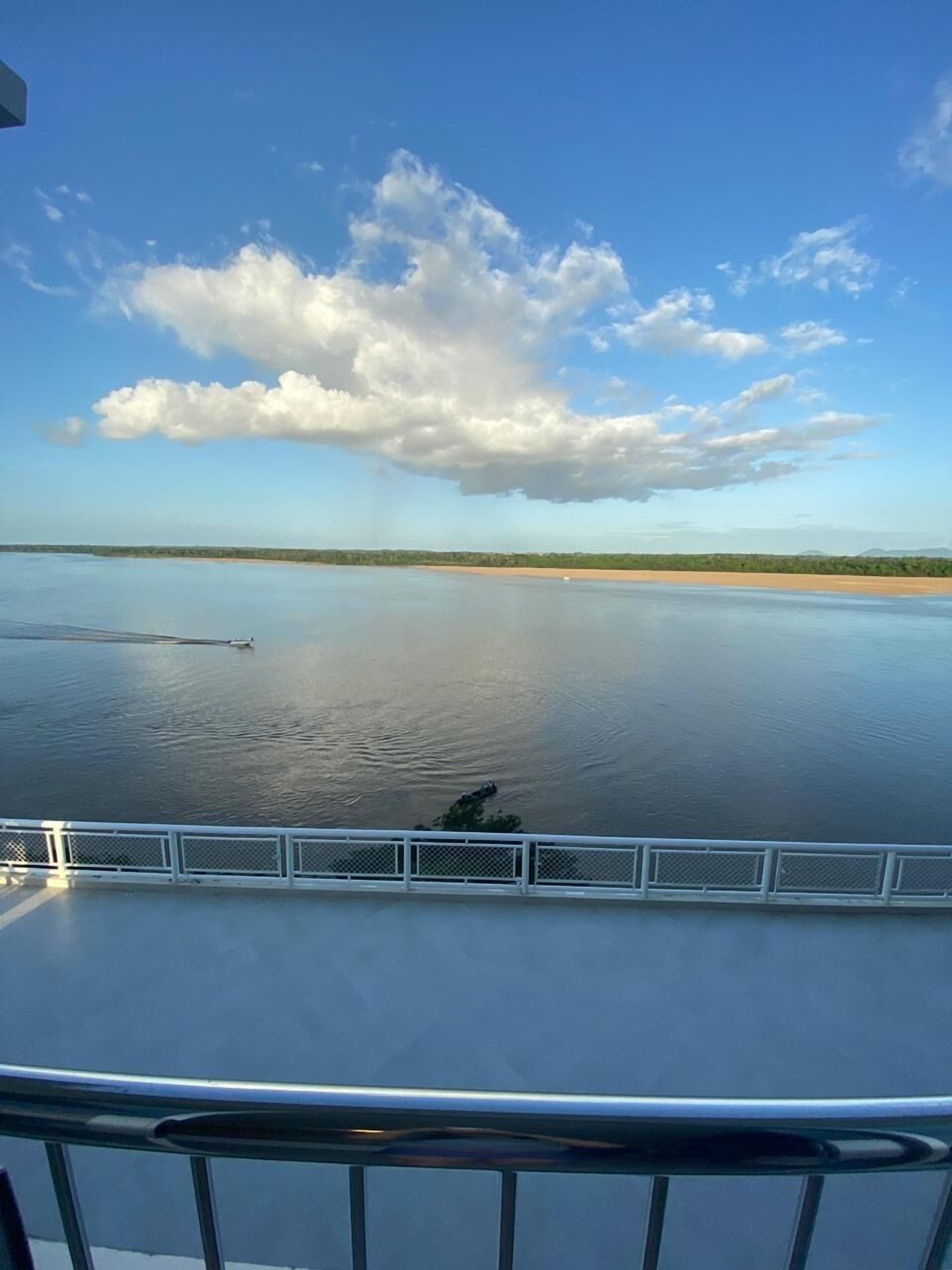 Una masa de agua tranquila con una costa lejana, cielo despejado y una barandilla blanca al frente.