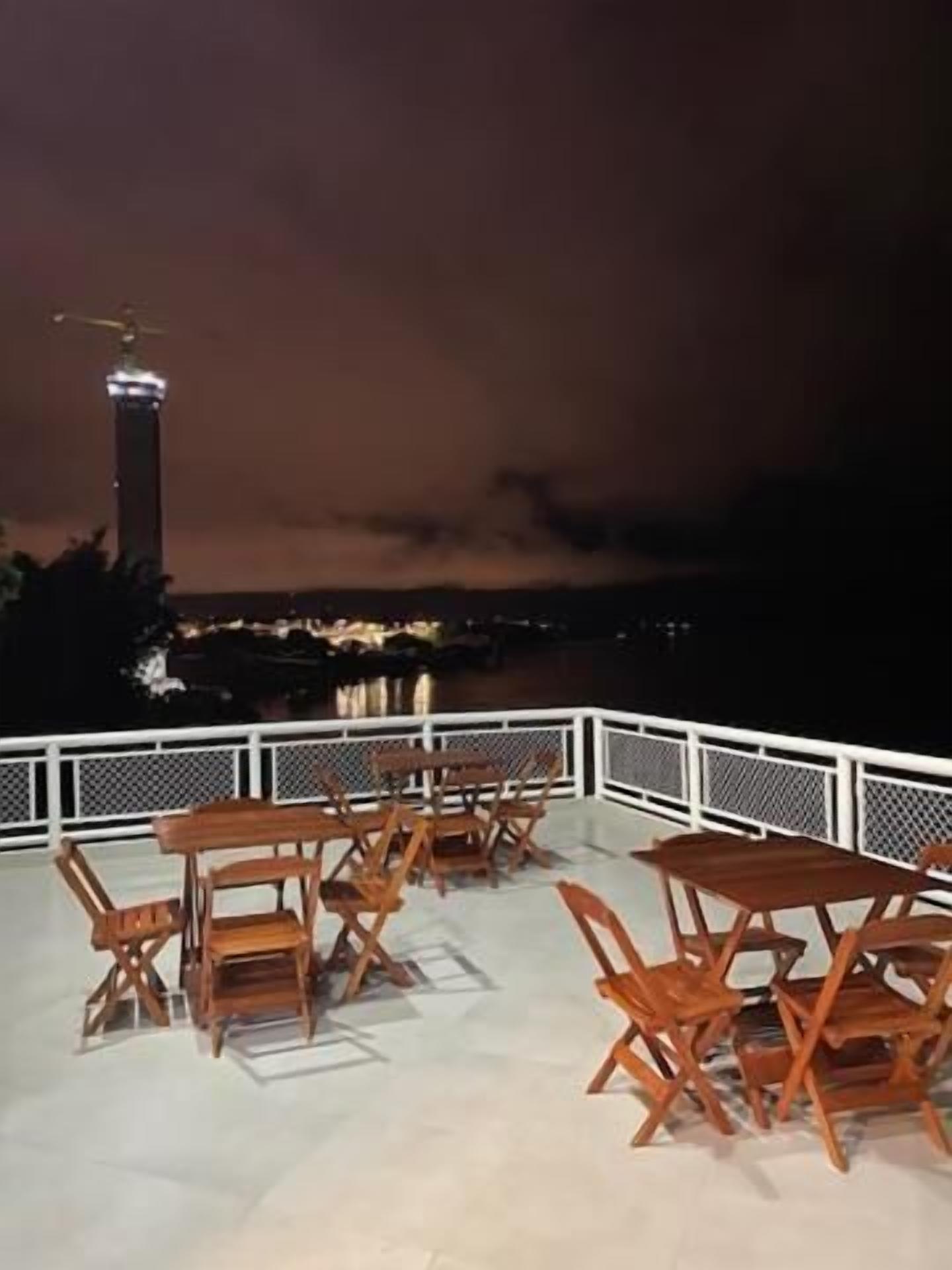 Un área de estar al aire libre con mesas y sillas de madera, barandilla blanca y vista a un paisaje urbano con un edificio alto a lo lejos.