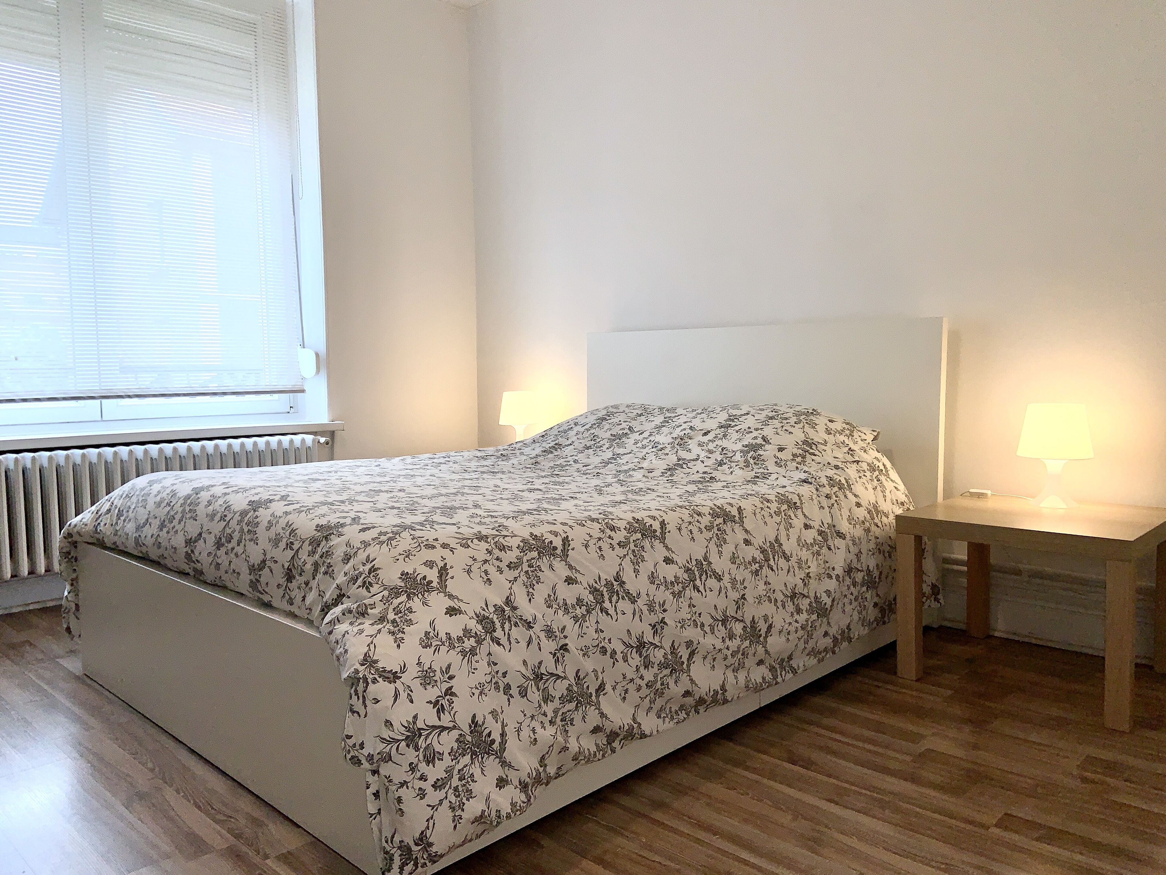 A bedroom with a bed, two bedside tables with lamps, a radiator, and a window with blinds.