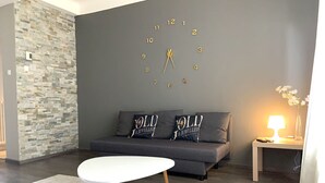 A living room with a stone wall, grey sofa, white table, and a clock on the wall. - Heart of Metz, 2 rooms free parking wifi 75m² "Le Saint Bernard (Metz)