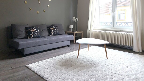 A modern living room with a grey sofa, a white round table, and a large wall clock. - Heart of Metz, 2 rooms free parking wifi 75m² "Le Saint Bernard (Metz)
