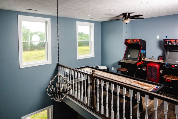 A room with a high ceiling, two windows, a chandelier, arcade games, and a staircase with a railing.