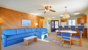 A living room with a blue sectional sofa, a wooden coffee table, and a dining area with a round table and blue chairs. - Sea Scape - Multiple Pools, Amenities, and Close to Beach! (Kitty Hawk)