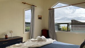 A bedroom with a bed, a bedside table with a mirror, a potted plant, and a window with curtains. - Moana Apartment with Bush Views and near Surf (Raglan)