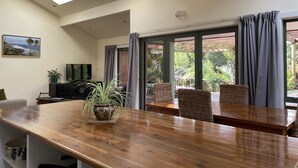 A spacious room with a wooden dining table, a television, and a potted plant. - Moana Apartment with Bush Views and near Surf (Raglan)