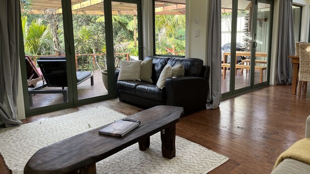 A spacious living area with a black leather sofa, a wooden coffee table, and a view of an outdoor deck with a wooden pergola and lush greenery. - Moana Apartment with Bush Views and near Surf (Raglan)