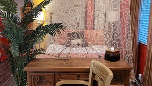 A wooden desk with drawers, a chair with a colorful cushion, a potted plant, and a patterned curtain. - Garden-front studio  (Phoenix)