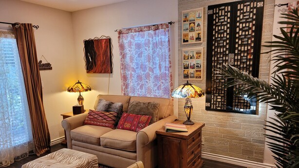 A cozy living room with a sofa, ottoman, and two lamps. A decorative wall panel and a potted plant are also visible. - Garden-front studio  (Phoenix)