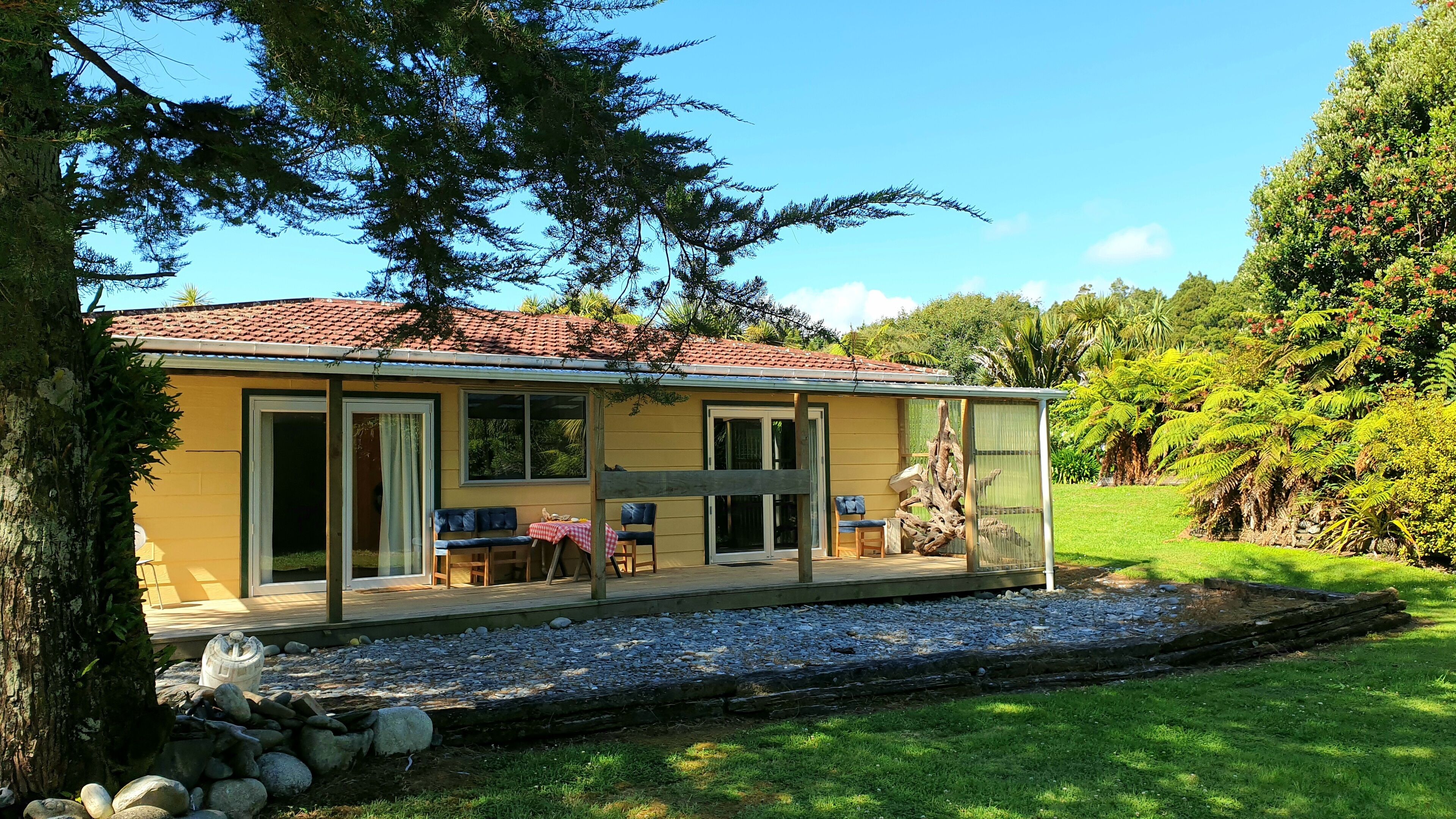 Una casa amarilla con techo de tejas rojas, amplios ventanales correderos y un patio con mobiliario exterior.