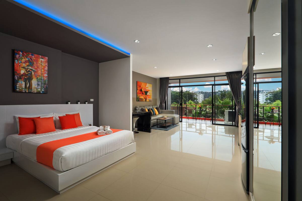 A modern hotel room with a large bed, a desk, and a sofa. There is a balcony with a view of the city.