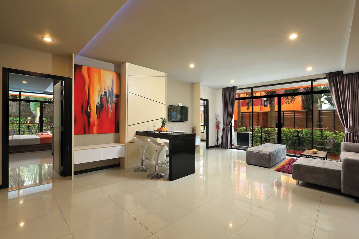 A modern living room with a black coffee table, a grey sofa, and a TV mounted on the wall. There is a large window with curtains and a view of the outdoors.