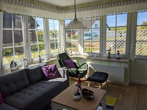 Et lyst rum med store vinduer, en grøn lænestol, en sofa og et lille bord med en flaske og glas.