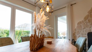 Una mesa de comedor de madera con un jarrón de plumas de plumeros, una lámpara geométrica y una pared de piedra al fondo.