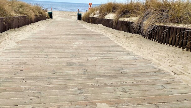 Pasarela de madera que lleva a la playa, bordeada por dunas de arena y un cartel que indica una profundidad de 10 metros.