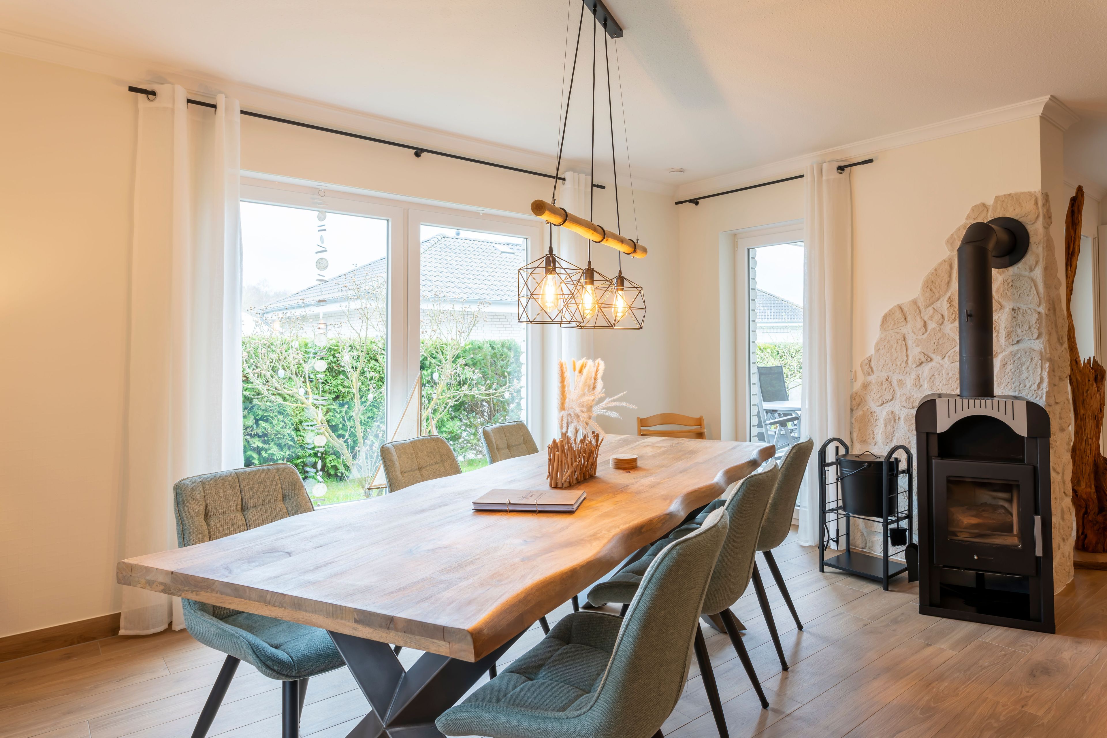 Een eetruimte met een grote houten tafel, stoelen, een hangende lamp en een stenen openhaard.