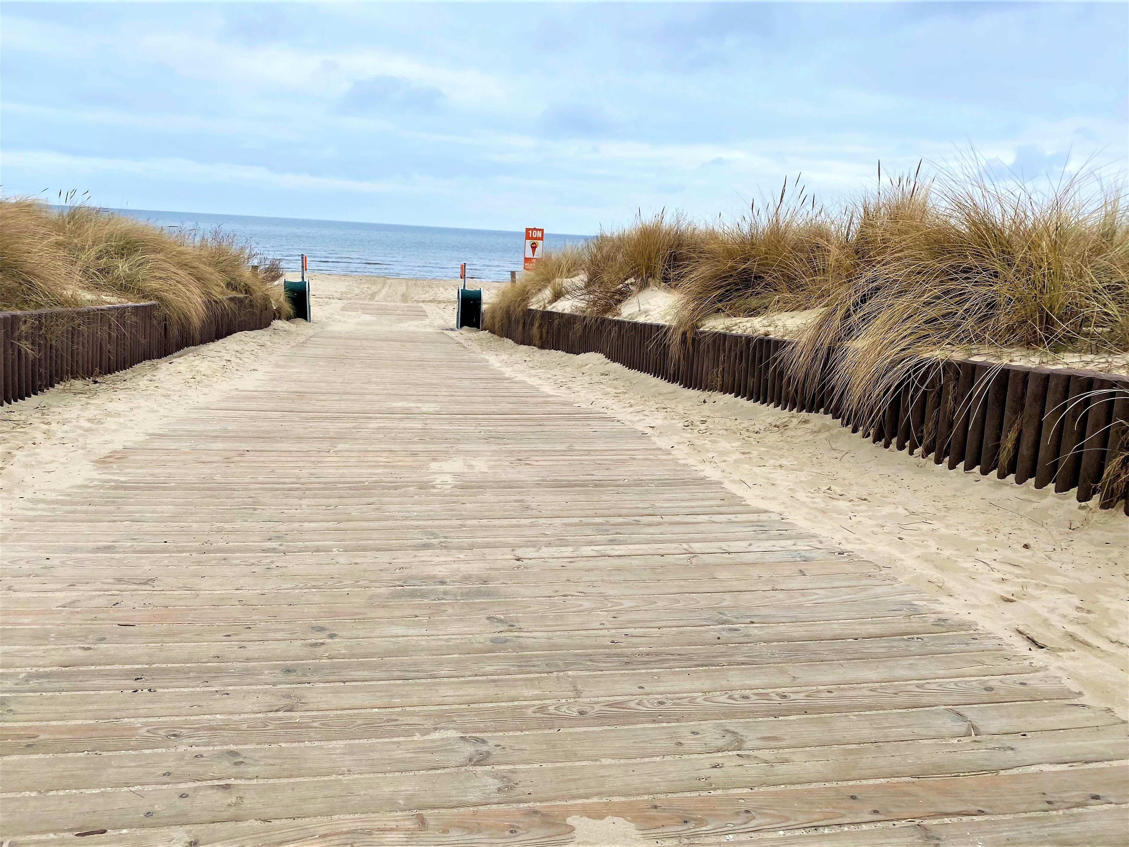 Pasarela de madera que lleva a la playa, bordeada por dunas de arena y un cartel que indica una profundidad de 10 metros.