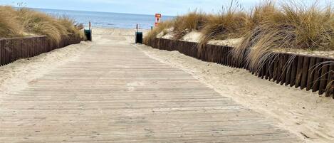 En træpromenade fører ned til stranden, stødt op af klitter og et skilt, der angiver en vanddybde på 10 meter.