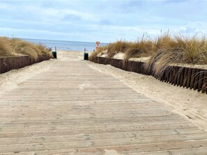 Une promenade en bois menant à la plage, bordée de dunes de sable et d’un panneau indiquant une profondeur de 10 mètres.