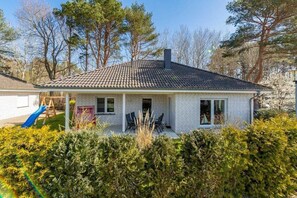 Une maison de plain-pied avec un toit en tuiles, une véranda abritée avec des chaises et un petit jardin agrémenté d’un toboggan.