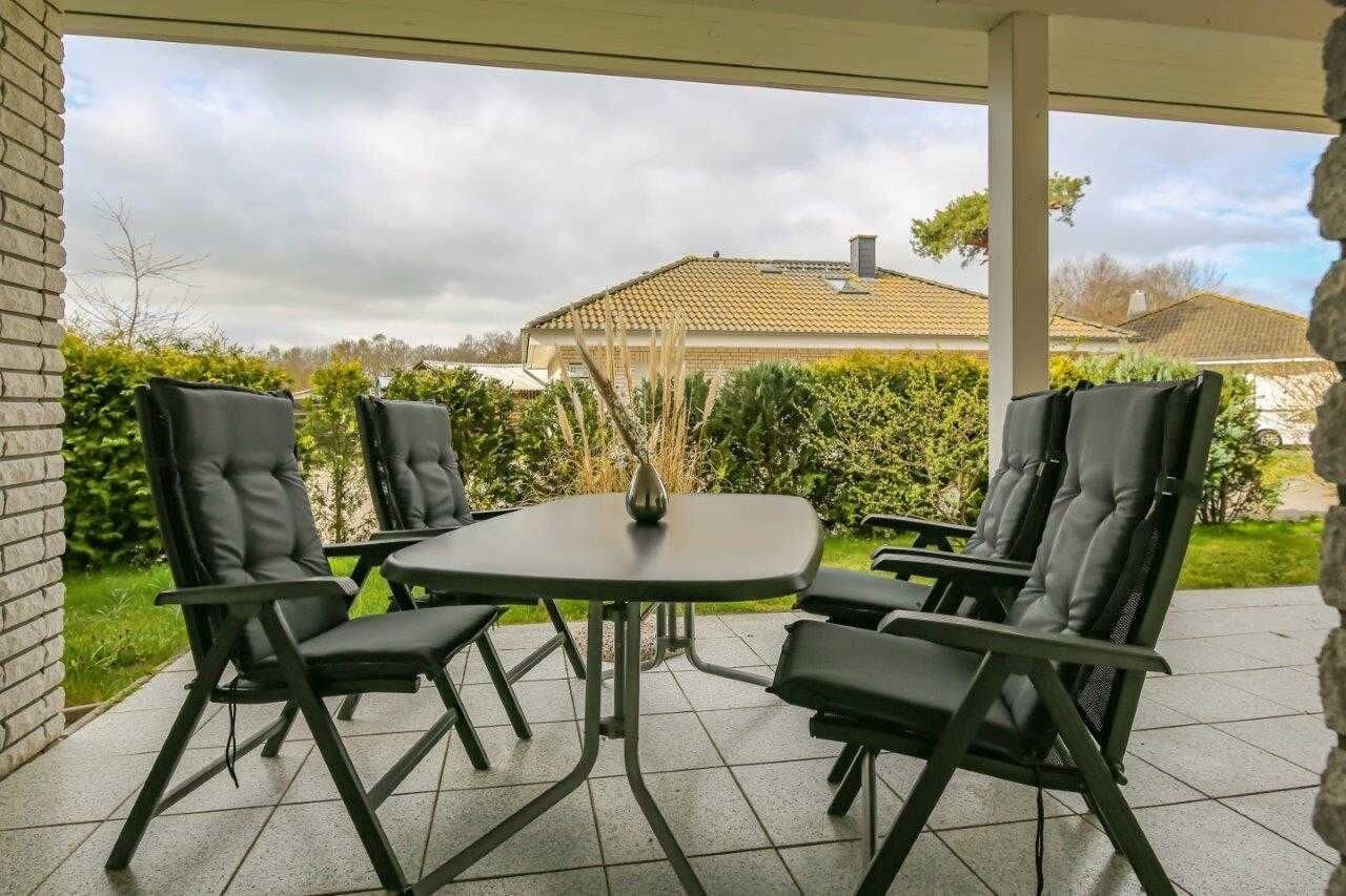 Une terrasse extérieure avec une table et quatre chaises, entourée de verdure et avec une maison en arrière-plan.
