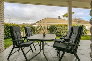 Eine Terrasse im Freien mit Tisch und vier Stühlen, umgeben von viel Grün und einem Haus im Hintergrund.