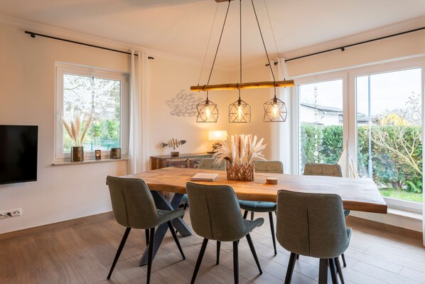 Un espace de salle à manger moderne avec une table en bois, quatre chaises, des luminaires suspendus et une vue sur la verdure à l’extérieur.