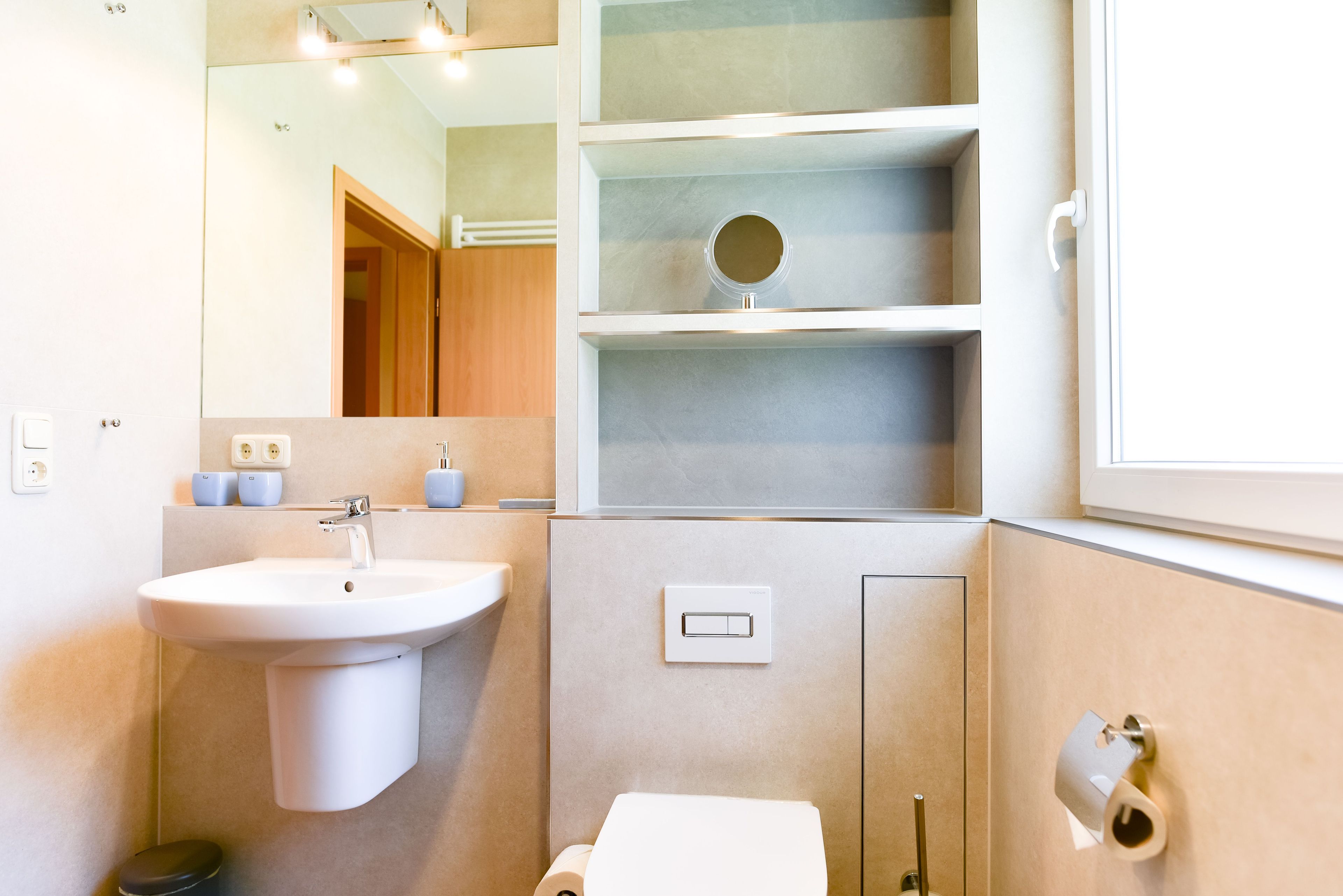 A modern bathroom with a sink, mirror, toilet, and shelves.