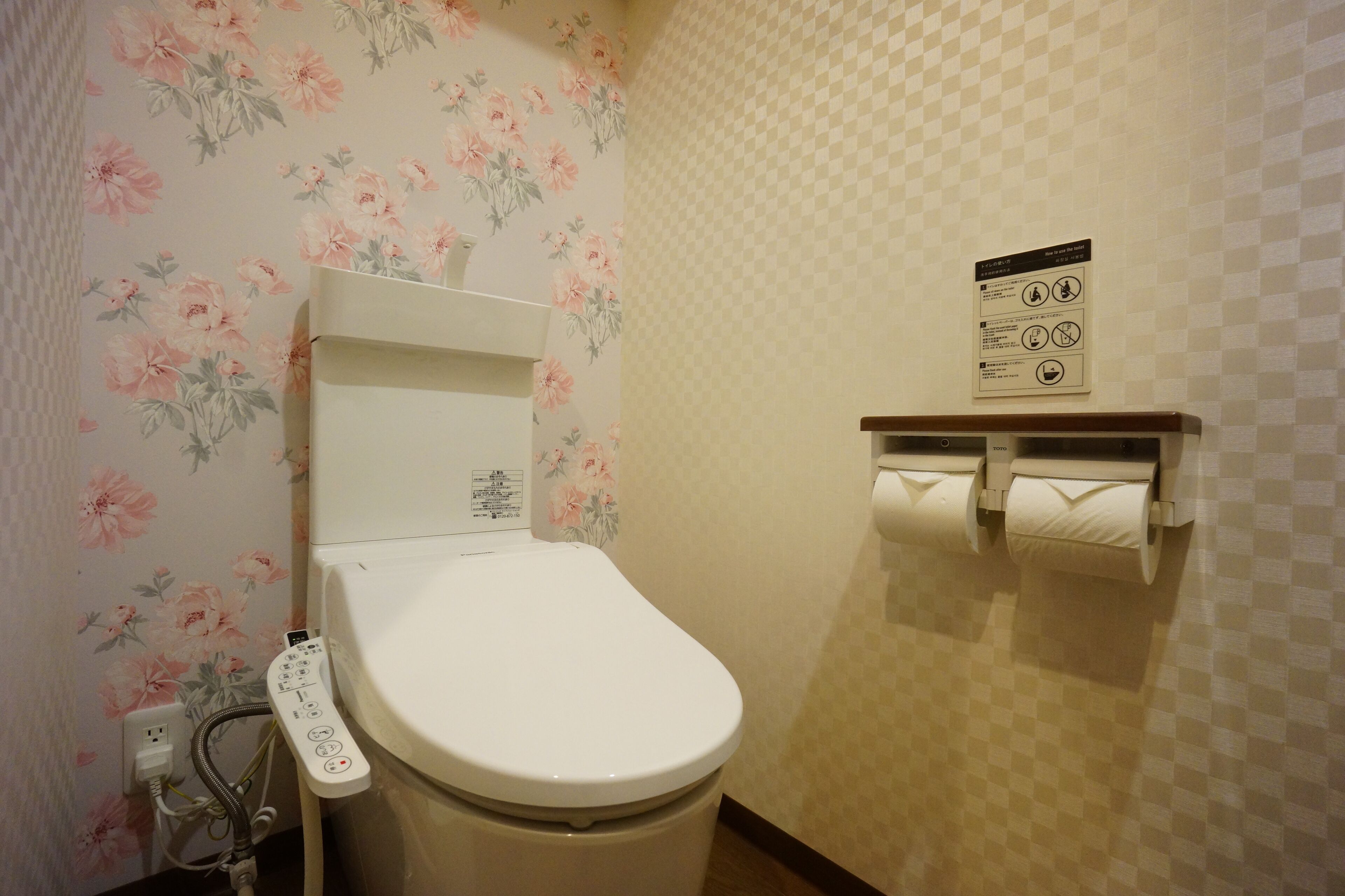 Des toilettes dotées d’un panneau de commande, d’un porte-rouleau de papier toilette et d’un papier peint à motifs floraux.
