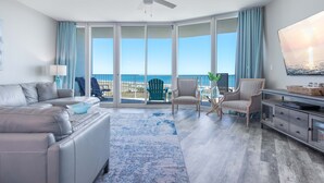A spacious living room with a large window, a ceiling fan, a sofa, a TV, and a dining area. - Timeless Three Bedroom Condo with Stunning Gulf Views! Complimentary Boat Slip! (Orange Beach)