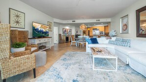 A spacious living room with a white sofa, a coffee table, and a television on a stand. There is a dining area with a chandelier and a kitchen in the background. - Beautifully Decorated Three Bedroom/Three Bath Condo Sleeping 10. Complimentary Boat Slip! (Orange Beach)