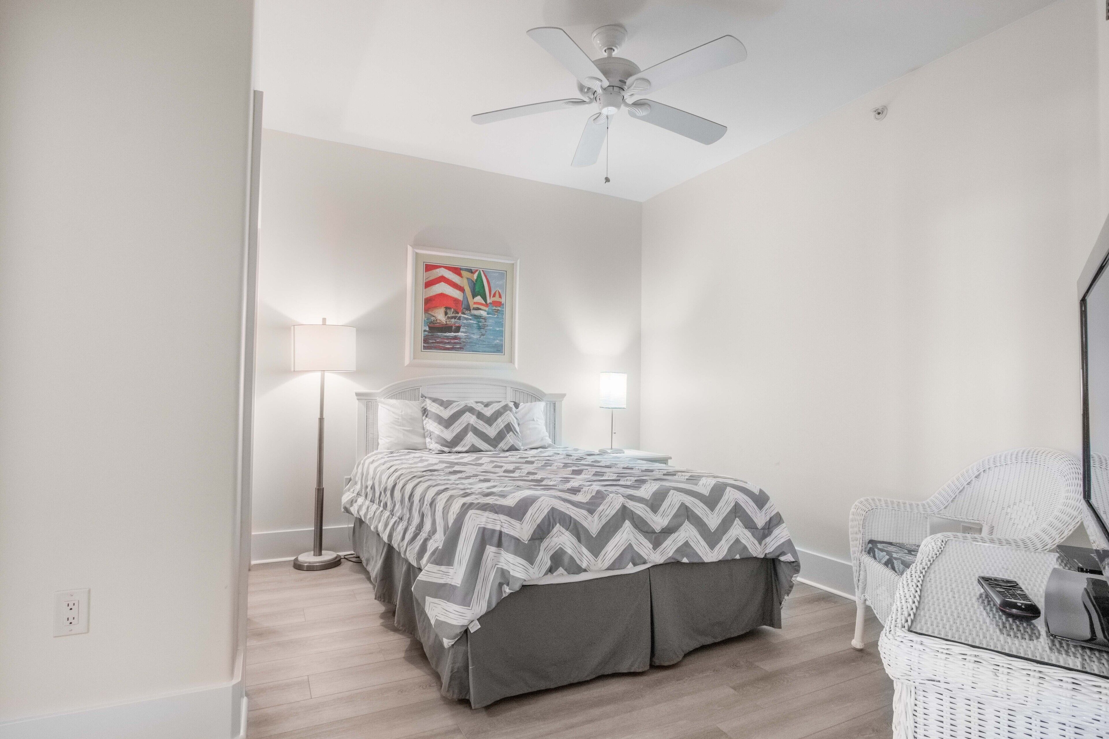 Une chambre à coucher avec un lit, une lampe sur pied, un ventilateur de plafond et une chaise en osier.