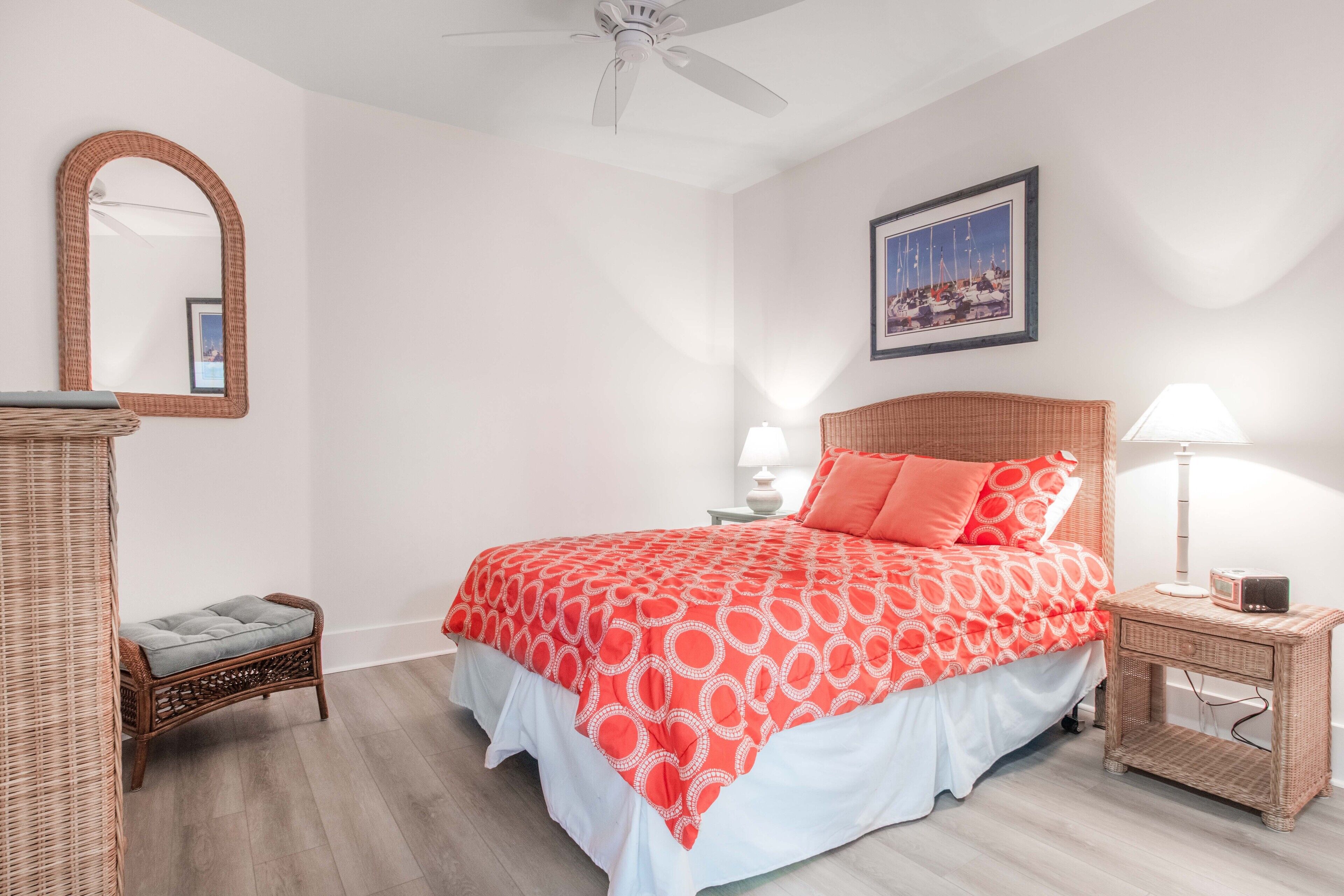 A bedroom with a bed, a nightstand, a lamp, a wicker chair, a mirror, and a framed picture on the wall.