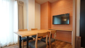 A modern dining area with a wooden table and chairs, a flat-screen TV mounted on the wall, and a large window with curtains. - WIRES HOTEL James zaka (Tokyo)