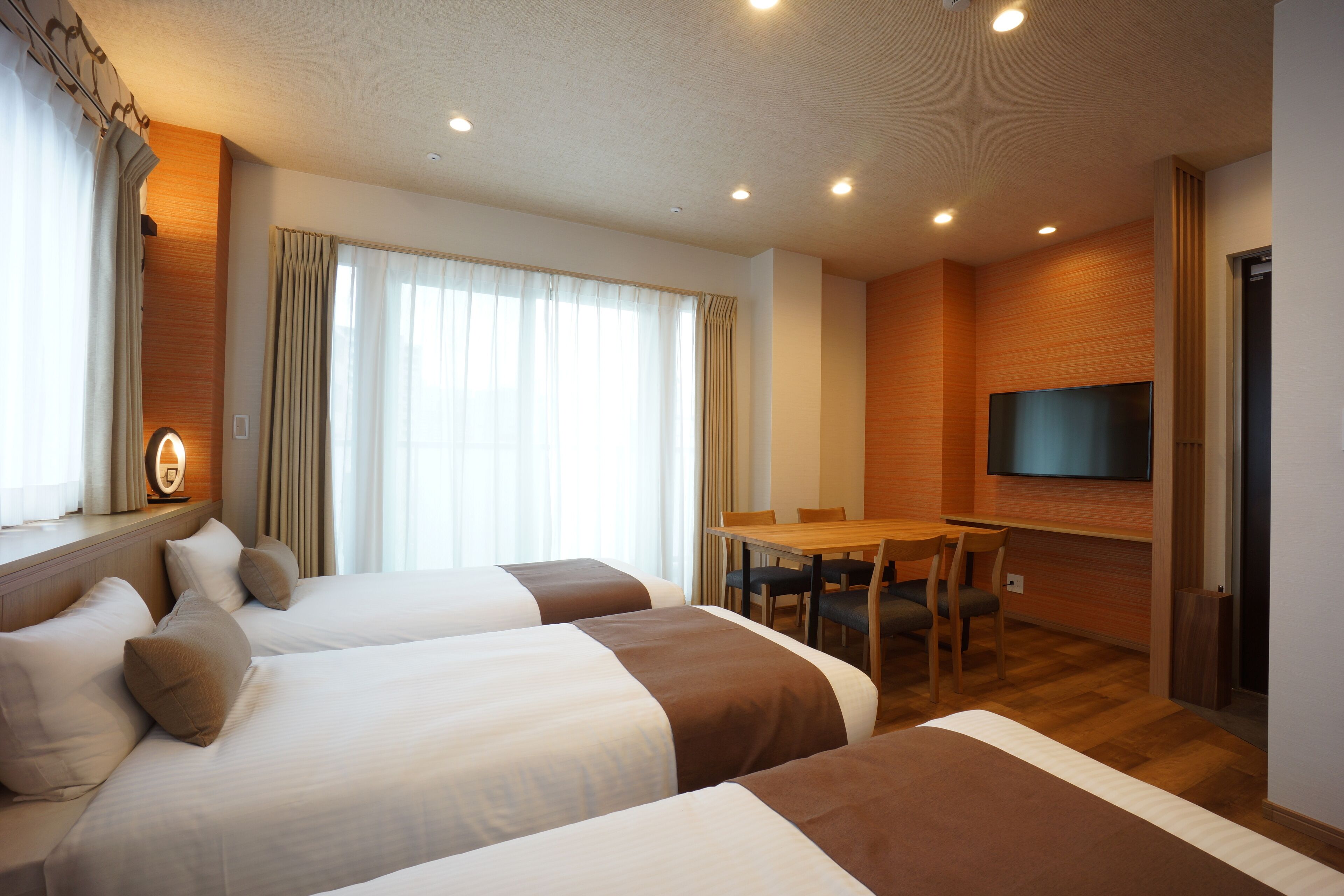 A hotel room with two beds, a dining table, chairs, a flat-screen TV, and a window with curtains.