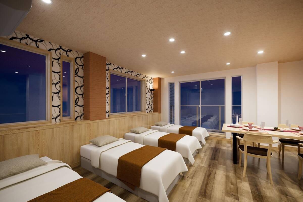 A hotel room with four single beds, a dining table with chairs, and large windows.