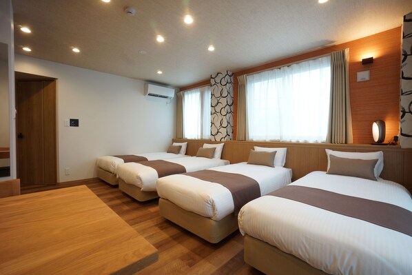 Habitación de hotel con cuatro camas dispuestas en fila, piso de madera, ventana grande con cortinas y aire acondicionado instalado en la pared.