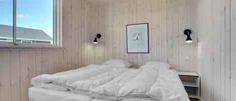 A bedroom with a wooden paneled wall, a bed with white bedding, two wall-mounted reading lamps, and a framed picture on the wall.