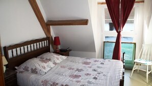 Un dormitorio con cabecera de madera, cama con ropa de cama floral, mesita de noche con lámpara roja y ventana con vistas al mar.