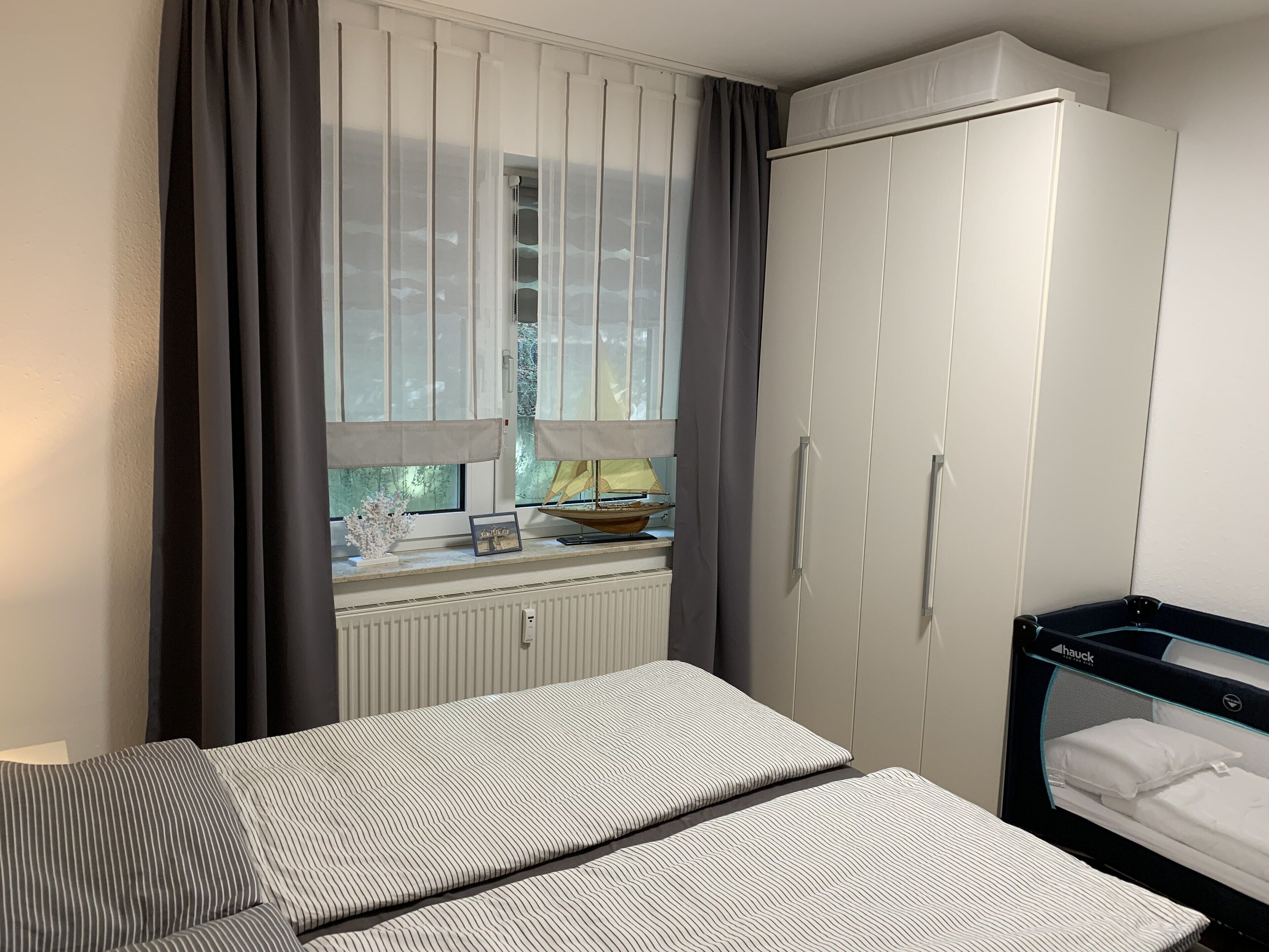 A bedroom with a bed, a wardrobe, a window with curtains, a radiator, and a small table with a lamp and picture frame.
